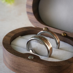 Puede incluir: Dos anillos de boda con bandas plateadas e incrustaciones de madera, presentados en un joyero de madera ovalado abierto, forrado con tela blanca. Los anillos son de la colección Timeless de Vestis Studios.