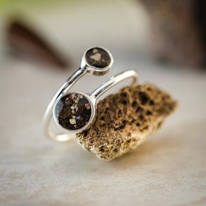 Puede incluir: Un anillo de plata con dos engastes circulares, cada uno con un material oscuro y texturizado. El anillo se muestra sobre una piedra rugosa y marrón. Se ven las palabras "VESTIS STUDIOS" y "TIMELESS COLLECTION".