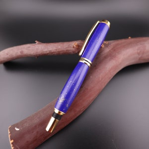 Puede incluir: Un bolígrafo azul y dorado con un diseño de lapislázuli. El bolígrafo está apoyado en una rama de madera marrón.