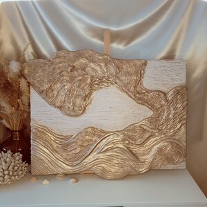 Könnte beinhalten: Ein strukturiertes abstraktes Gemälde mit goldener und weißer Farbe. Das Gemälde hat einen 3D-Effekt und erinnert an Wellen oder Berge.