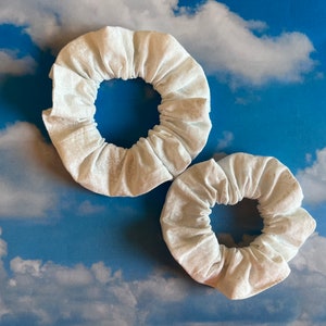 May include: Two white fabric scrunchies, one larger than the other, against a blue sky with white clouds background.