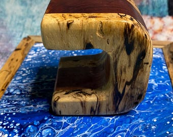 May include: A handcrafted wooden phone stand with a distinctive, curved design. The stand has a dark brown stripe across the top and is made of light-coloured wood with dark grain patterns. The stand is displayed on a blue and white surface.