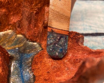 May include: A rectangular wooden pendant with a leather cord. The top portion is light brown wood, transitioning to a dark blue and green resin base. The pendant is displayed against a textured, reddish-brown wooden background.