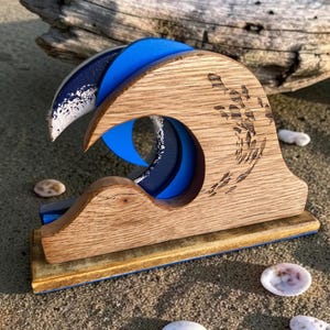 Puede incluir: Escultura de madera con forma de ola con detalles en azul y blanco. El diseño de la ola presenta un efecto de capas con un interior azul y una cresta azul oscuro y blanca. La base es rectangular con un borde azul. Sobre una superficie arenosa.