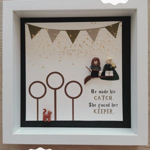 Könnte beinhalten: Ein gerahmtes DIY-Geschenk mit weißem Rahmen, mit einem schwarzen Hintergrund mit einem Design zum Thema Quidditch. Enthält zwei Figuren, Besen und den Text "He made his CATCH. She found her KEEPER."