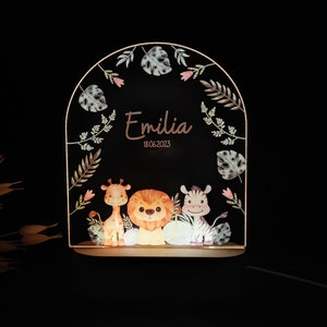 May include: A wooden night light with a whimsical jungle theme. The light features a giraffe, lion, and zebra with a leafy border. The name "Emilia" and the date "18.06.2023" are inscribed on the light.