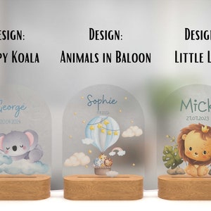 May include: Three personalised night lights with different designs. The first features a sleeping koala, the second has animals in a hot air balloon, and the third has a lion. Each light has a wooden base and a clear acrylic panel with a name and date.