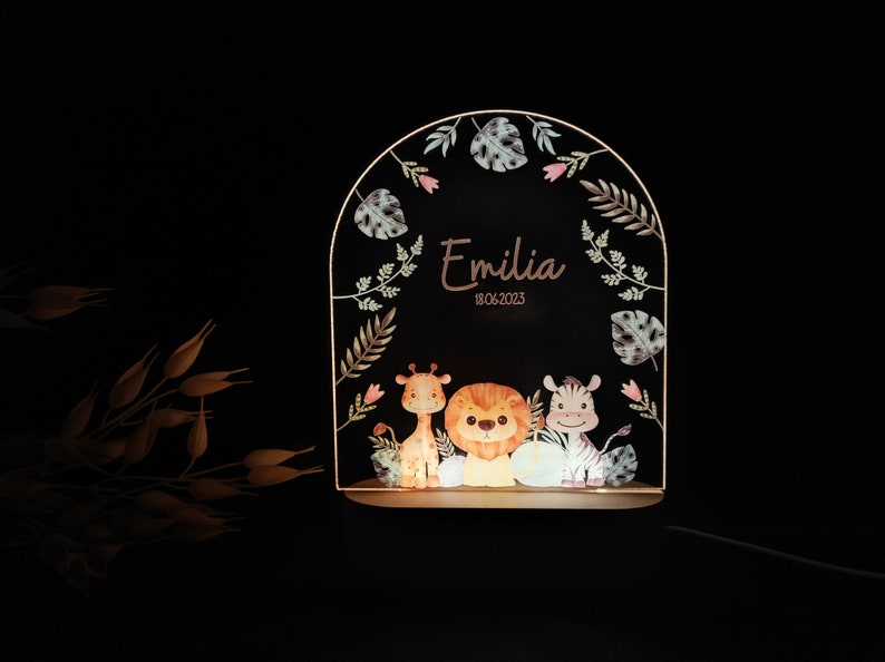 May include: A wooden night light with a whimsical jungle theme. The light features a giraffe, lion, and zebra with a leafy border. The name "Emilia" and the date "18.06.2023" are inscribed on the light.