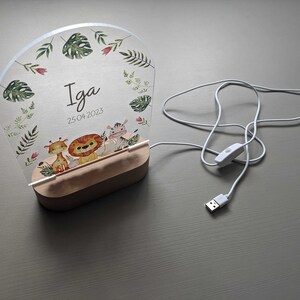 May include: A white acrylic night light with a wooden base. The light features a jungle animal theme with a giraffe, lion, and zebra. The light is personalised with the name "Iga" and the date "25.04.2023".