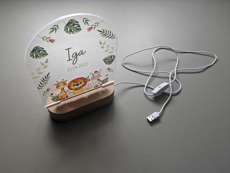 May include: A white acrylic night light with a wooden base. The light features a jungle animal theme with a giraffe, lion, and zebra. The light is personalised with the name "Iga" and the date "25.04.2023".