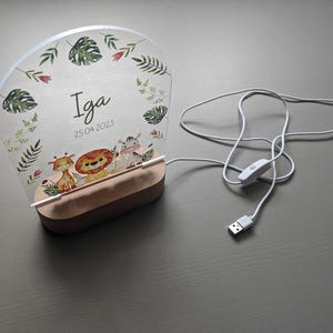 Può includere: Una luce notturna in acrilico bianco con un tema giungla. La luce presenta un design a foglia con un leone, una giraffa e una zebra. La luce è personalizzata con il nome "Iga" e la data "25.04.2023".