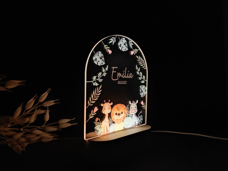 May include: A wooden arch-shaped night light with a clear acrylic panel featuring a watercolour illustration of a giraffe, lion, and zebra with a floral border. The name "Emilia" and the date "08.06.2023" are printed on the panel.