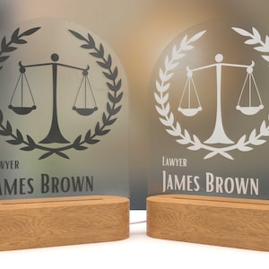 Könnte beinhalten: Zwei beleuchtete Anwaltsschilder mit einem Justitia-Emblem und dem Namen "James Brown". Die Schilder sind auf klarem Acryl geätzt und stehen auf Holzsockeln. Der Text "LAWYER" steht über dem Namen.