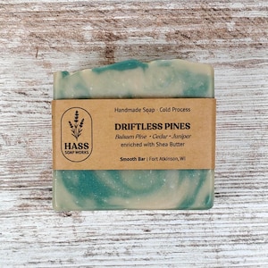 May include: A handmade soap bar with a marbled teal and white design. The bar is wrapped with a brown paper label that reads "DRIFTLESS PINES" and lists ingredients. The soap is from Hass Soap Works.