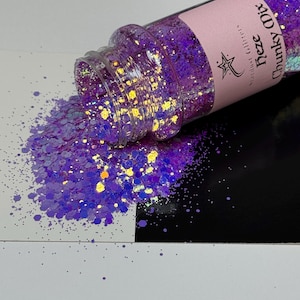 May include: A clear plastic bottle of purple glitter, labeled "Chunky Mix Heze Stardust Glitter," is tipped over, spilling glitter onto a white and black surface. The glitter is a mix of purple and iridescent colors, creating a sparkling effect.