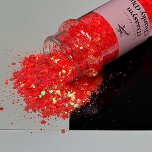 May include: A clear glass jar of chunky, neon orange glitter is tipped over, spilling onto a black and white surface. The glitter is a mix of shapes and sizes, reflecting light. The jar's label reads "Daasym Chunky Mix".