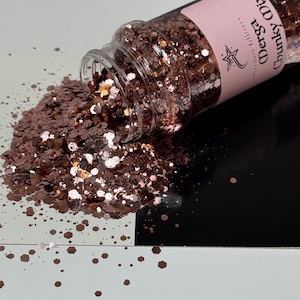 May include: A clear jar of glitter is tipped over, spilling out a mix of copper and rose gold glitter. The glitter is in various shapes and sizes, including hexagonal and round. The jar has a pink label with the text "Derga Funky Mix".