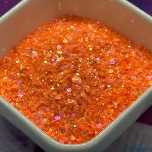 May include: A close-up of a small white bowl filled with orange glitter. The glitter has a holographic finish, with iridescent pink and gold flecks.