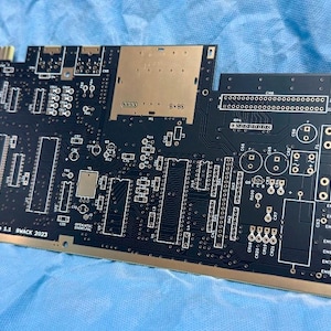 Könnte beinhalten: Eine schwarze Leiterplatte mit goldfarbenen Kontaktpunkten und verschiedenen Bestückungsflächen für elektronische Bauteile. Die Platine ist mit weißem Text beschriftet, darunter PCB ASSY NO.250468, PCB NO.252011 REV.A, Replica 1.1 und BWACK 2023.