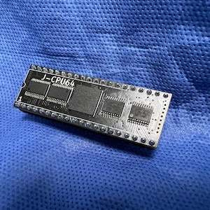 May include: A dark grey rectangular circuit board, featuring the text "J-CPU64". It has several black rectangular components and silver pins. The background is a textured blue fabric. The board is a component of electronic devices.