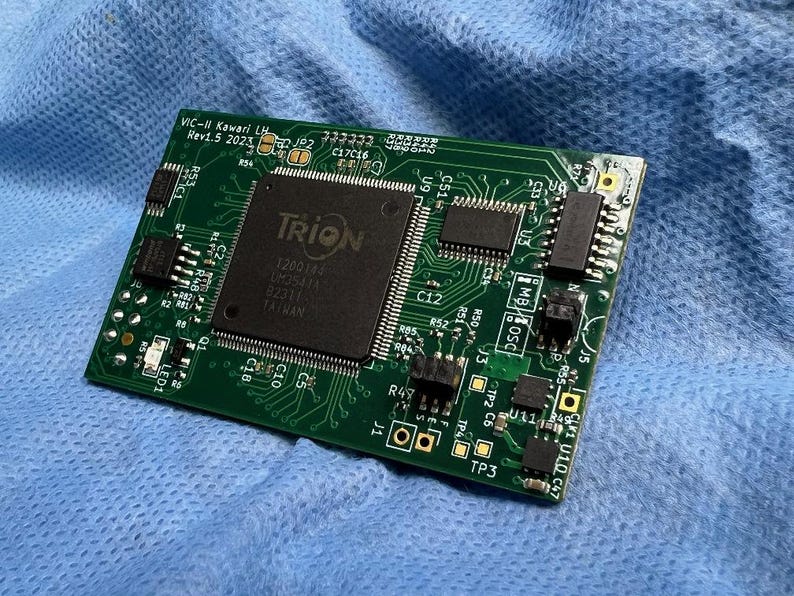 Op de afbeelding: Een groene printplaat met de tekst "VIC-II Kawari LH Rev1.5 2023" en een groot zwart vierkant met "TRION". De printplaat heeft diverse elektronische componenten en ligt op blauwe stof.