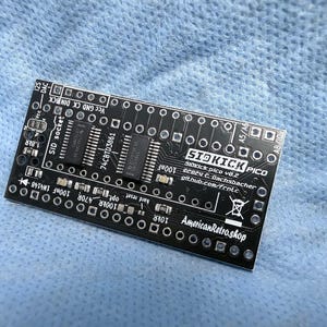 May include: Black circuit board with the text "SIOKICK PICO" and "AmericanRetro.shop". The board has various electronic components, including chips and resistors, and is set against a blue background.