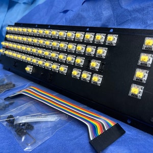 May include: A black keyboard plate with a grid of yellow mechanical keyboard switches. The plate is connected to a rainbow ribbon cable.