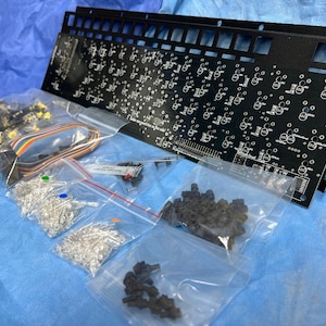 Könnte beinhalten: Eine schwarze Leiterplatte mit mehreren Reihen kleiner, runder Bauteile. Die Platine ist von mehreren durchsichtigen Plastiktüten umgeben, die verschiedene kleine elektronische Bauteile enthalten, darunter LEDs, Widerstände und Drähte.