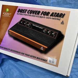May include: A white box with a graphic of an Atari 2600 VCS console. The box reads "Dust Cover for Atari" and "High Quality Accessories for Your Retro Machines." The console is black with wood-grain accents and silver switches.