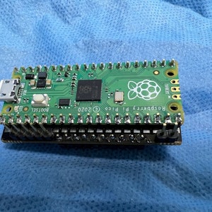 May include: A green Raspberry Pi Pico microcontroller board with a USB-C port, a button, and a Raspberry Pi logo. The board has a black header with gold pins.