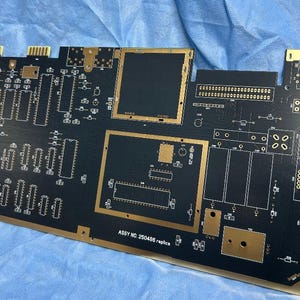 Peut inclure: Une carte de circuit imprimé noire avec des traces et des composants dorés. La carte comporte diverses sections rectangulaires et carrées, avec le texte "ASSY NO. 250408 replica" imprimé dessus. La carte est sur une surface bleue.