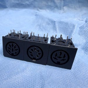 Könnte beinhalten: Drei schwarze elektronische Steckverbinder mit mehreren Pins. Jeder Stecker hat einen kreisförmigen Anschluss mit mehreren Stiftlöchern. Die Stecker sind nebeneinander auf einer blauen Oberfläche angeordnet.