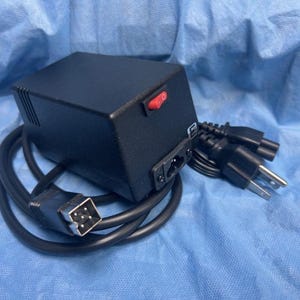 Puede incluir: Un dispositivo electrónico negro con un interruptor de encendido rojo, un cable de alimentación y múltiples clavijas. El dispositivo tiene forma rectangular y está conectado a un cable negro con un conector de cuatro pines.