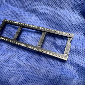 Op de afbeelding: Een zwart plastic onderdeel met talrijke kleine, zilverkleurige metalen oogjes in rijen gerangschikt. Het onderdeel heeft een rechthoekige vorm met drie open secties. Het ligt op een blauwe, getextureerde stof.