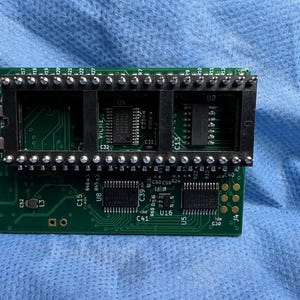 Op de afbeelding: Een groene printplaat met meerdere zwarte connectoren en ge&iuml;ntegreerde circuits. De printplaat heeft verschillende componenten die zijn gelabeld met letters en cijfers, waaronder "U2", "C32" en "U16". De achtergrond is een blauwe stof.