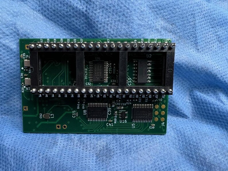 Op de afbeelding: Een groene printplaat met meerdere zwarte connectoren en ge&iuml;ntegreerde circuits. De printplaat heeft verschillende componenten die zijn gelabeld met letters en cijfers, waaronder "U2", "C32" en "U16". De achtergrond is een blauwe stof.