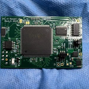 Op de afbeelding: Een groene printplaat met diverse elektronische componenten, waaronder een grote centrale chip en kleinere ge&iuml;ntegreerde circuits. De tekst "VIC-II Kawari LH Rev1.5 2023" is zichtbaar. De printplaat ligt op een blauw oppervlak.