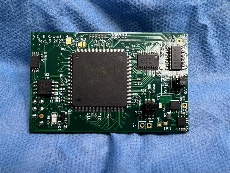 Op de afbeelding: Een groene printplaat met diverse elektronische componenten, waaronder een grote centrale chip en kleinere ge&iuml;ntegreerde circuits. De tekst "VIC-II Kawari LH Rev1.5 2023" is zichtbaar. De printplaat ligt op een blauw oppervlak.
