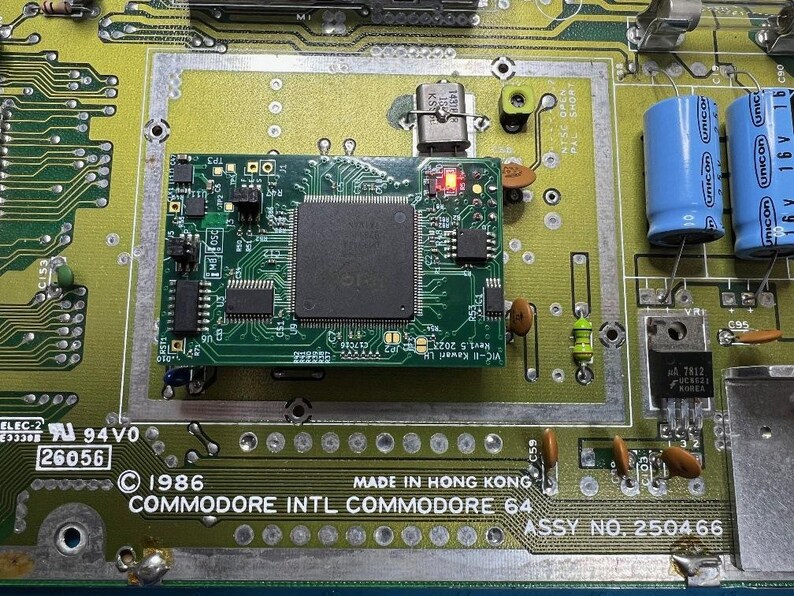Op de afbeelding: Close-up van een Commodore 64 printplaat. De groene printplaat bevat diverse elektronische componenten, waaronder chips, condensatoren en weerstanden. De tekst "COMMODORE INTL COMMODORE 64" is zichtbaar, samen met "MADE IN HONG KONG" en het jaar 1986.