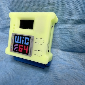 May include: A neon yellow electronic device with a black screen and a colourful label that reads "WiC 64". The device has several small indicator lights and a blue connector on the bottom. The background is a light blue fabric.