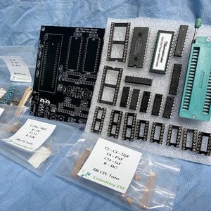 Può includere: Una collezione di componenti elettronici per un tester CPU Z80, tra cui una scheda a circuito stampato nera, vari chip, zoccoli e un componente verde. Diversi componenti sono confezionati in sacchetti di plastica trasparente con etichette.