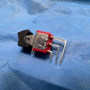 Puede incluir: Un interruptor basculante eléctrico negro y rojo con clavijas plateadas. El interruptor tiene una palanca negra y una base roja. El interruptor está sobre una superficie azul.
