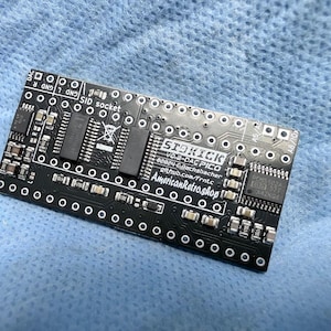 May include: A black circuit board featuring electronic components like chips and connectors. The board is labelled with "STOKICK" and "VO.2-DAC PICO", and the website "AmericanRetro.shop". It rests on a blue surface. The board is covered in various electronic components.