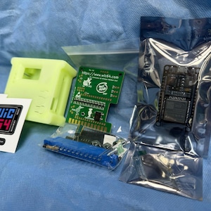Peut inclure: Ensemble de composants électroniques, comprenant une carte de circuit imprimé verte, une carte de circuit imprimé noire dans un sac scellé, un boîtier en plastique jaune, des autocollants avec le texte "WIC 64" et d'autres petites pièces. Les composants sont disposés sur une surface bleue.