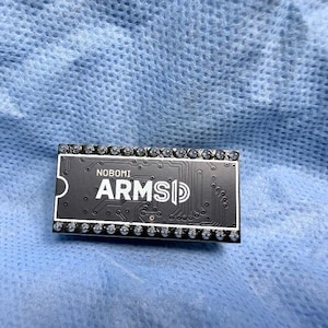 May include: A black circuit board with the text "NOBOMI ARMSD" in white. The board is rectangular with a series of small, silver-colored components along the edges. The background is a light blue, textured fabric.
