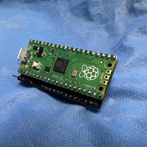 Puede incluir: Una placa de microcontrolador Raspberry Pi Pico verde con un puerto micro USB y un cabezal con pines chapados en oro. La placa tiene el texto "Raspberry Pi Pico" impreso en ella.
