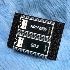 Könnte beinhalten: Zwei schwarze, rechteckige elektronische Leiterplatten mit weißem Text und weißen Anschlüssen, montiert auf einem Stück schwarzem Schaumstoff. Die obere Platine ist mit ARM2SD und die untere mit SD2 beschriftet, beide tragen den Namen NOBOMI.