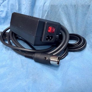 Peut inclure: Un bloc d'alimentation rectangulaire noir doté d'un interrupteur à bascule rouge lumineux et d'un port d'entrée à deux broches. L'appareil comprend un long câble noir se terminant par un connecteur métallique circulaire à trois broches internes.