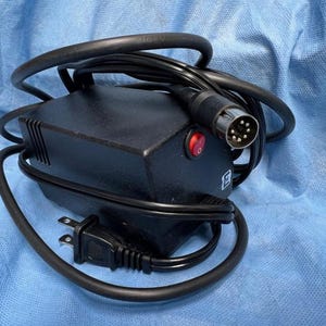May include: A black electronic device with a power cord, a red power button, and a multi-pin connector. The device has a rectangular shape and is on a blue surface.