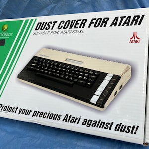 May include: A white box with green stripes on the side. The box has a picture of a beige Atari 800XL computer with a black keyboard. The text on the box reads "DUST COVER FOR ATARI" and "SUITABLE FOR: ATARI 800XL". The Atari logo is in the upper right corner of the box. The text "Protect your precious Atari against dust!" is at the bottom of the box.
